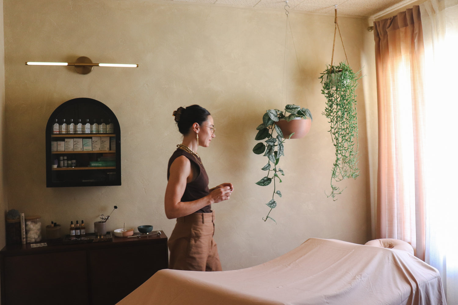 A woman in a spa room stands beside a massage table, looking toward hanging plants and a window. The room has warm beige walls, a wall-mounted shelf with bottles, and soft natural light from the right.