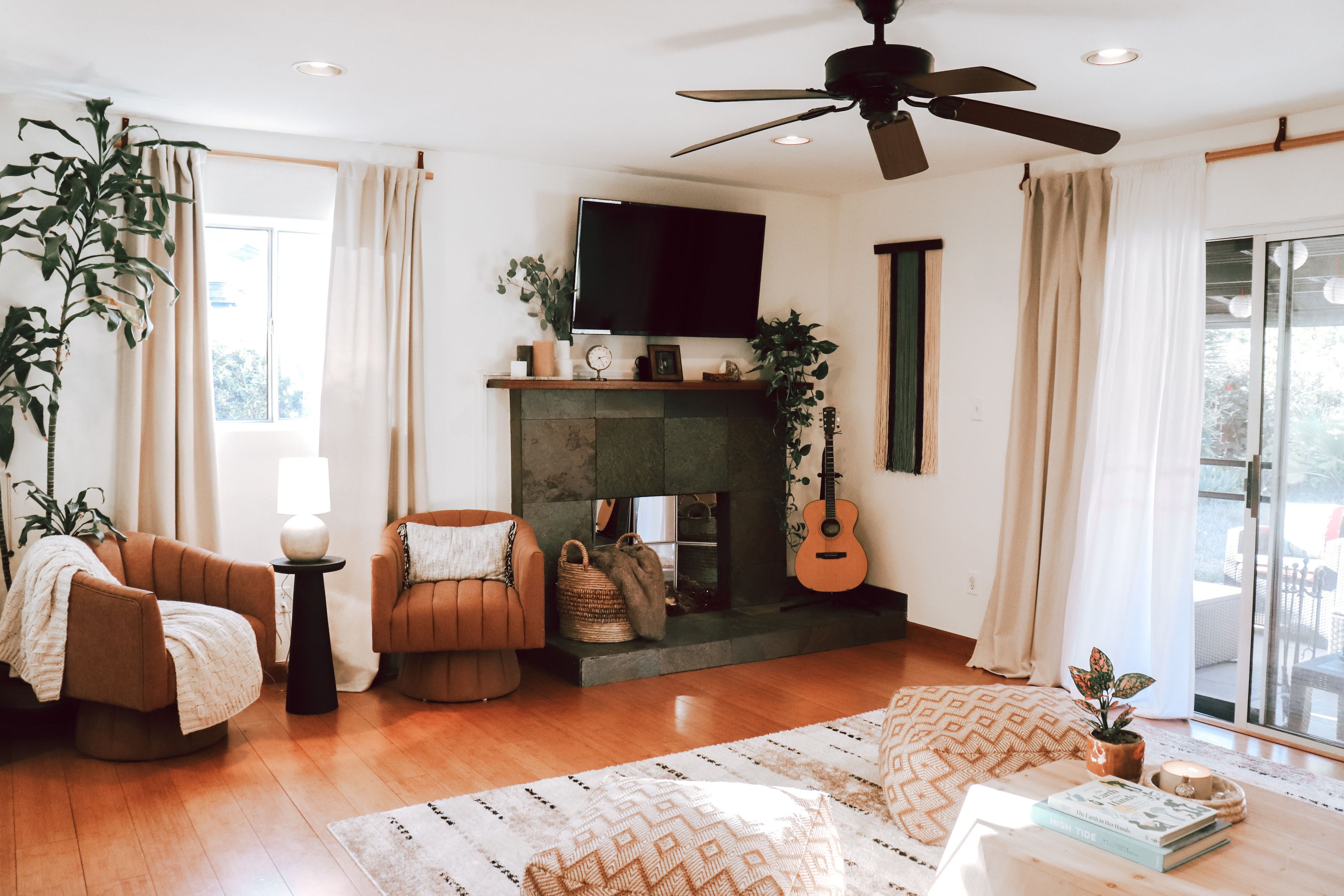 A bright, modern living room with warm wood floors, a stone fireplace, two brown armchairs, a patterned area rug, and a ceiling fan.