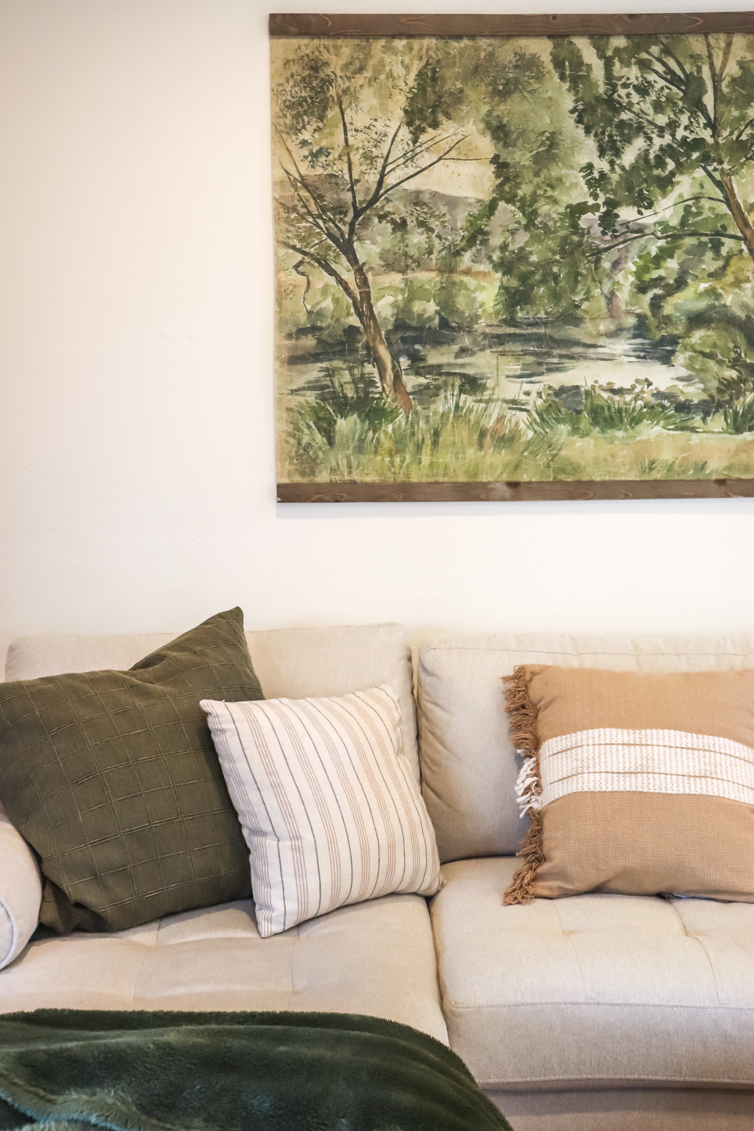 A framed landscape tapestry hanging on a white wall above a beige sofa with green and striped throw pillows.