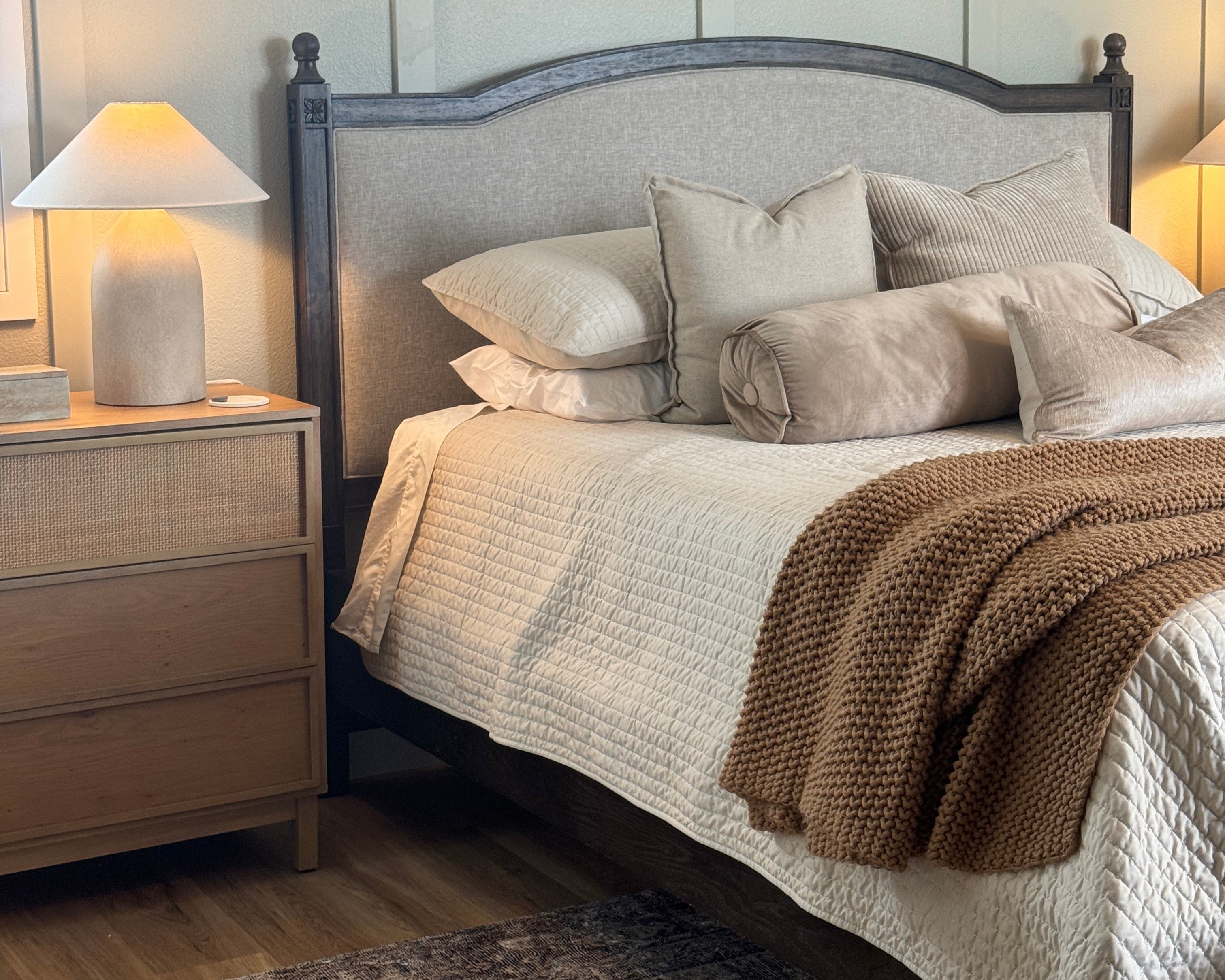 A bed with a dark wood frame and gray upholstered headboard, dressed in cream bedding with multiple pillows and a brown knit throw blanket, shown in a bedroom setting.