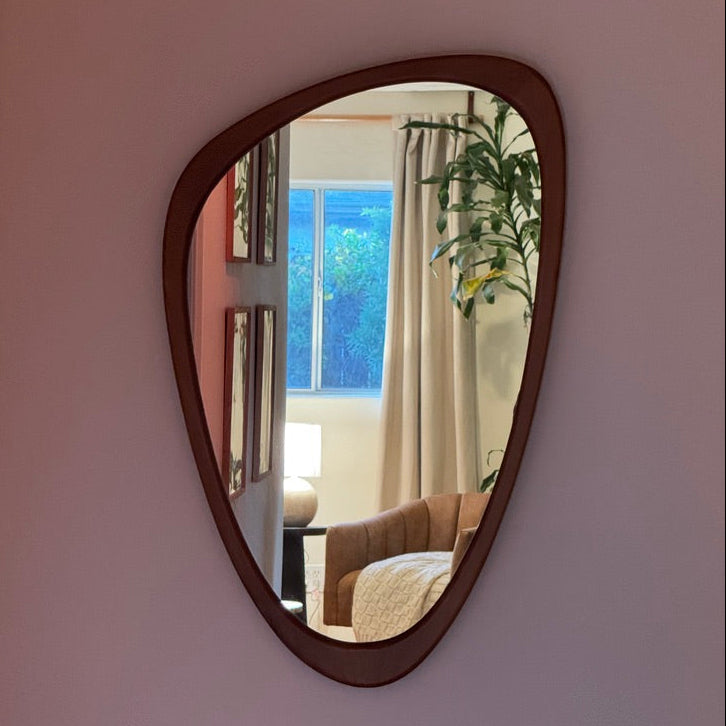 An organic-shaped wall mirror with a dark brown frame mounted on a light pink wall, reflecting a cozy bedroom interior with a window, curtains, and a plant.