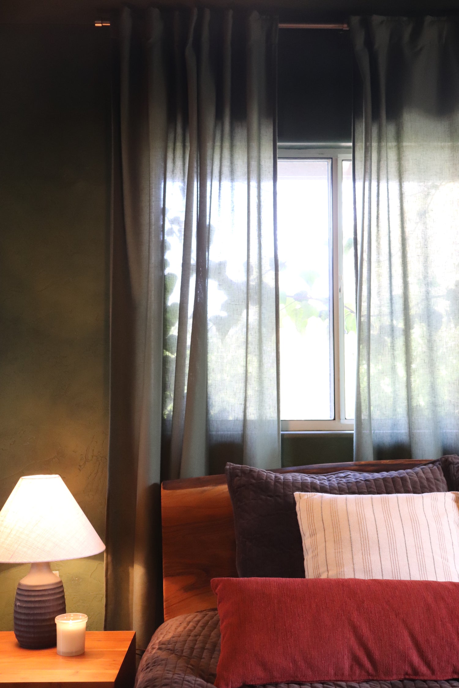 A bedroom scene showing dark gray curtains hanging over a window, with part of a bed featuring red and striped bedding, a bedside lamp, and a wooden nightstand visible.