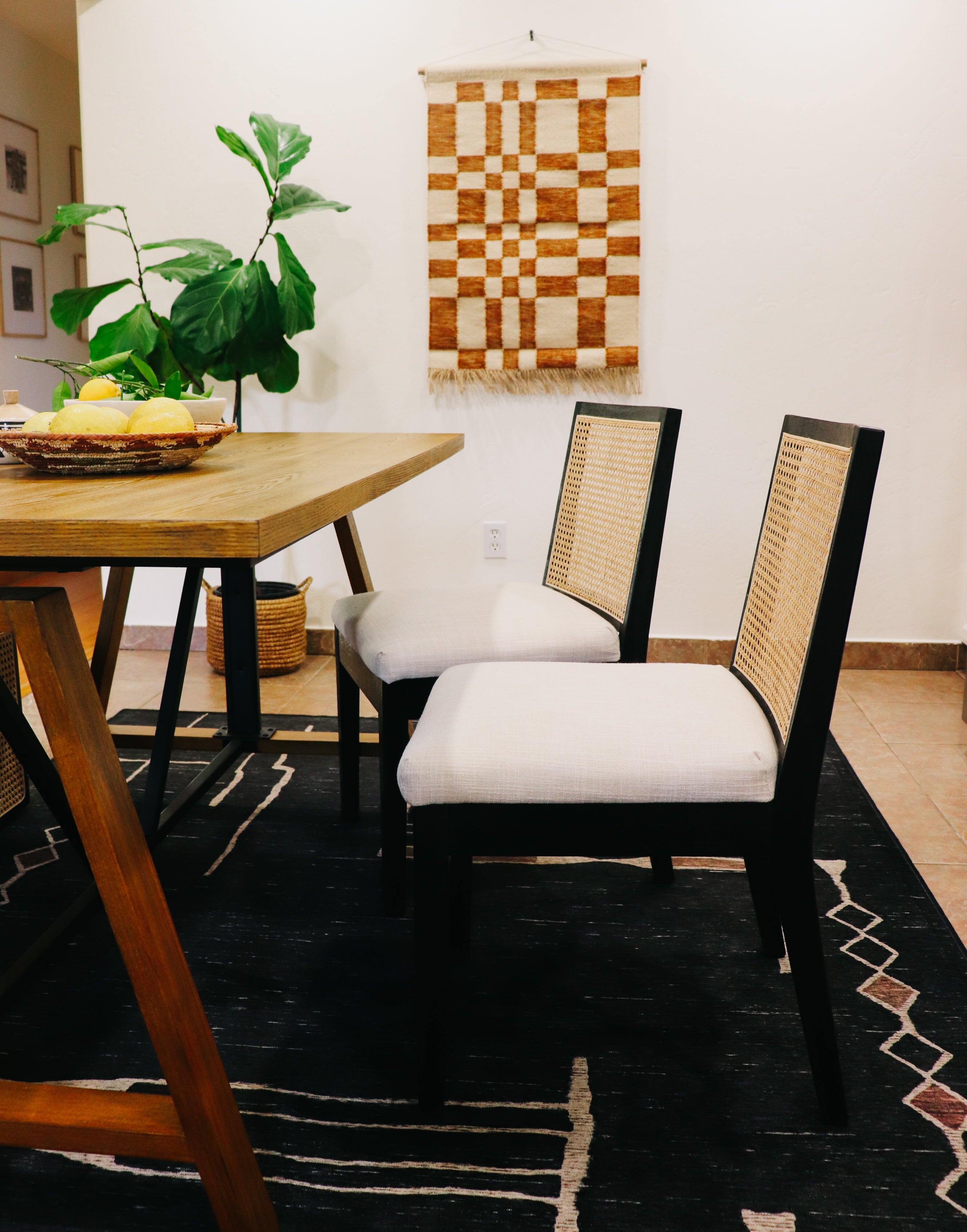 A dining room scene showing two black chairs with white cushions and woven cane backs, positioned around a wooden table on a black patterned rug. A green plant and a woven wall hanging are visible in the background.