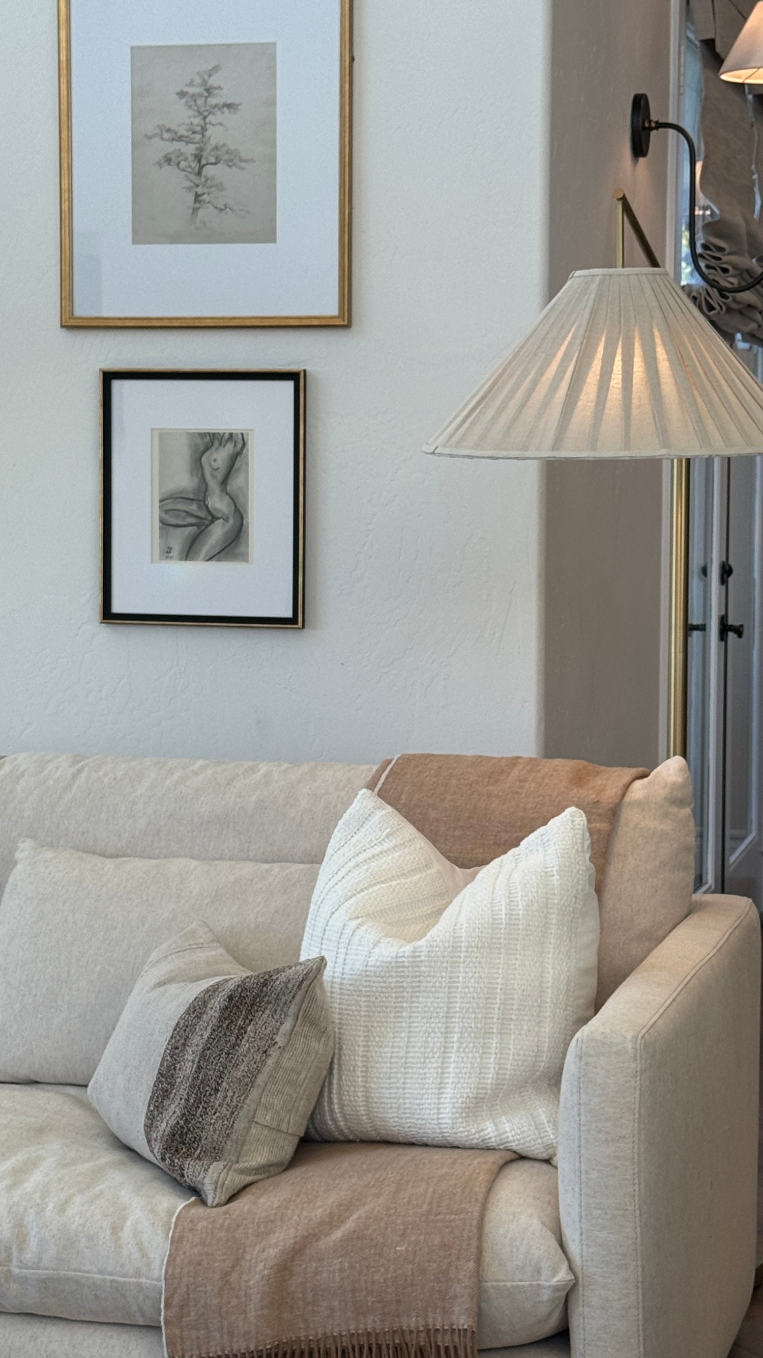 A cozy living room corner featuring a light beige sofa adorned with textured cream, white, and gray throw pillows and a beige throw blanket. Above the sofa, two framed minimalist art prints hang on a textured off-white wall, alongside a black wall lamp and a pleated white floor lamp.