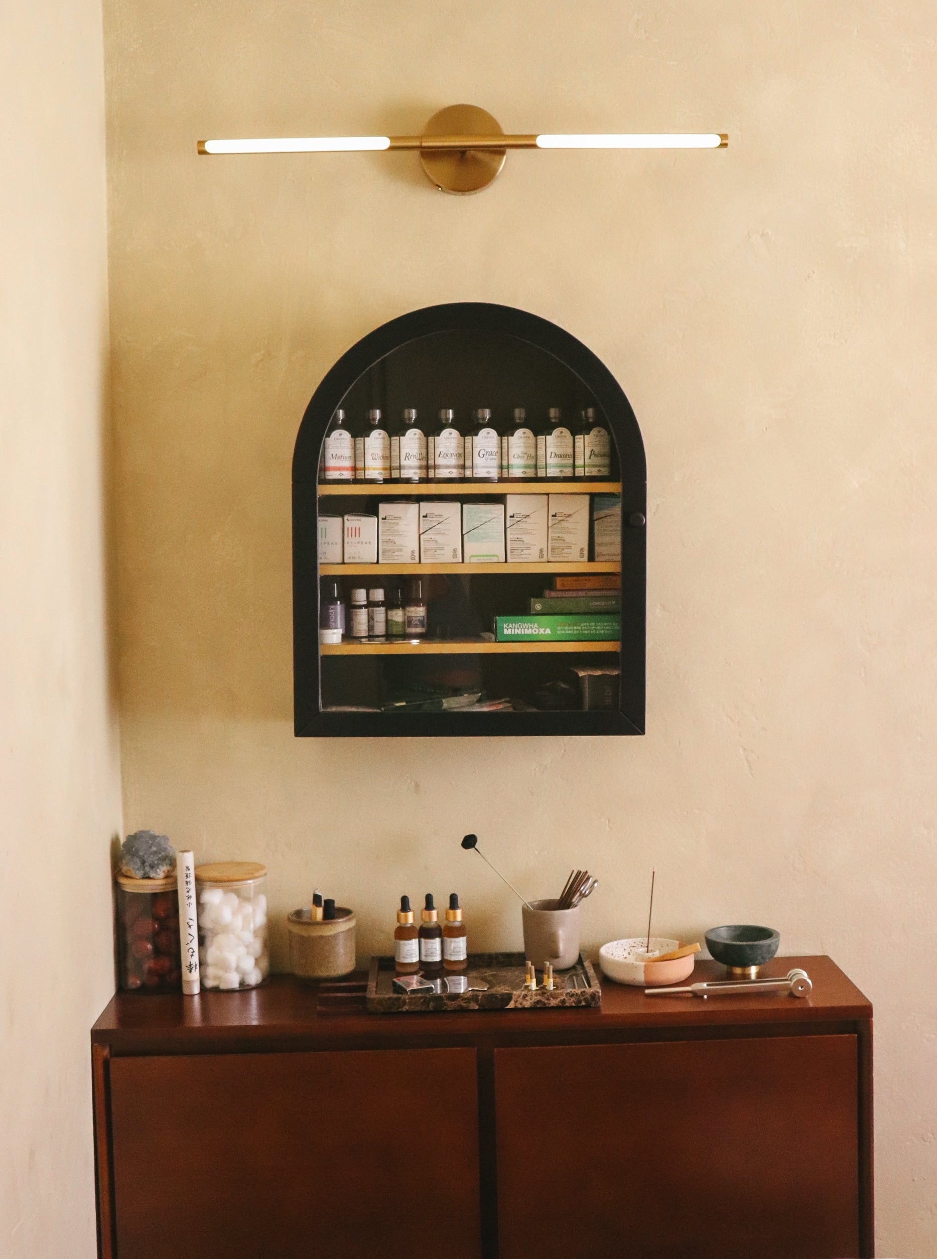 A warm-toned interior scene with a dark wood cabinet against a beige wall, a black arched wall-mounted shelf filled with bottles, and a modern horizontal bar light above. No person or animal is present, but this is not a clean isolated product shot; it shows the product in a styled real environment, so subject_type is Lifestyle.