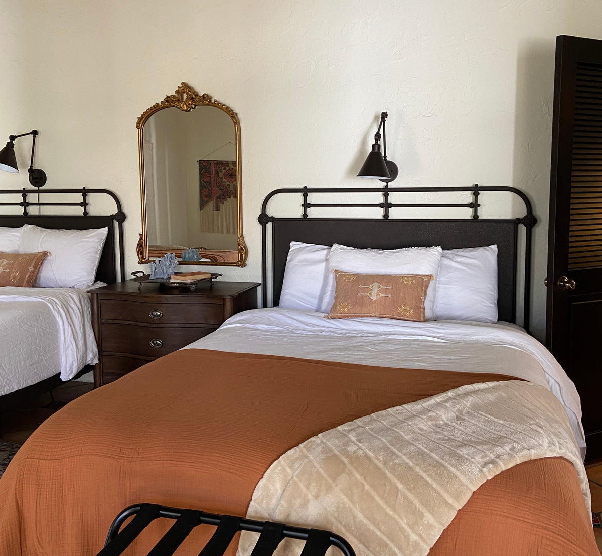 A bedroom scene showing a bed with a rust-orange comforter, white bedding, a cream throw blanket, and a black metal frame. A matching bed is partially visible below, along with a wall-mounted mirror, bedside table, and wall sconce.