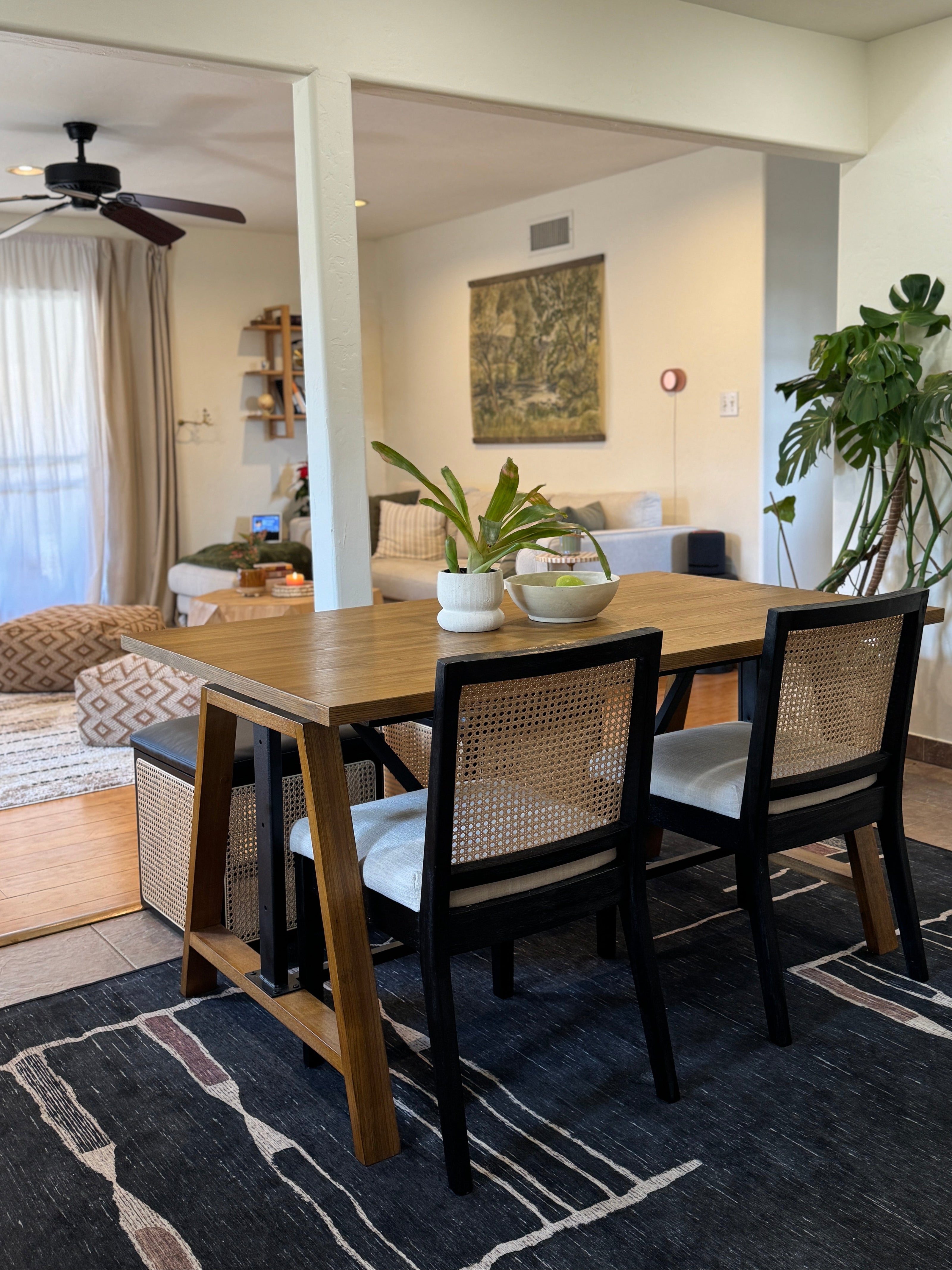 A modern dining table with a light wood top and black legs, paired with four black chairs featuring woven cane backs and white seat cushions, set on a dark patterned rug in a bright, open-plan home.