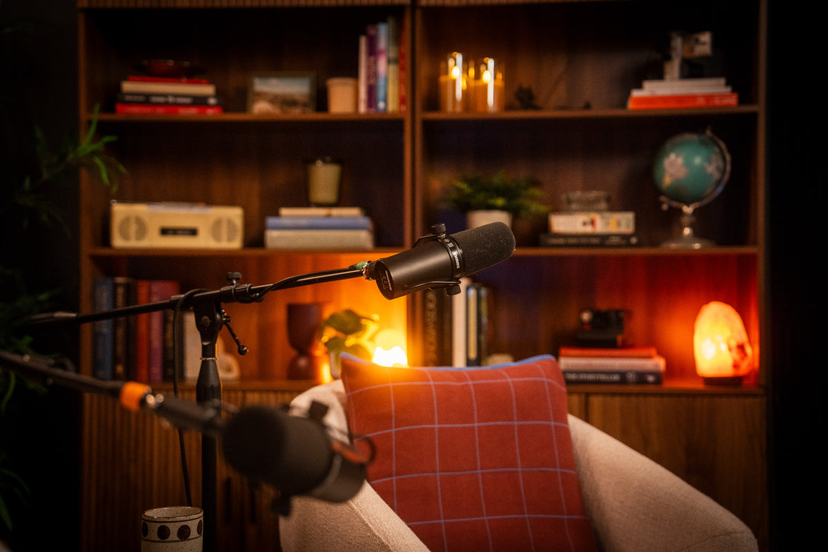 A professional studio setup featuring two black microphones on adjustable boom arms positioned in front of a cozy, warmly lit bookshelf with a red plaid cushion in the foreground.