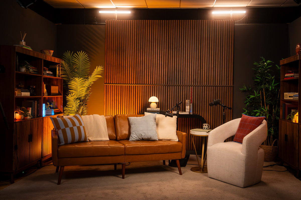 A styled interior scene with a brown leather sofa, cream and striped throw pillows, a white armchair with a red pillow, shelving, plants, and warm ambient lighting against a slatted wood wall.
