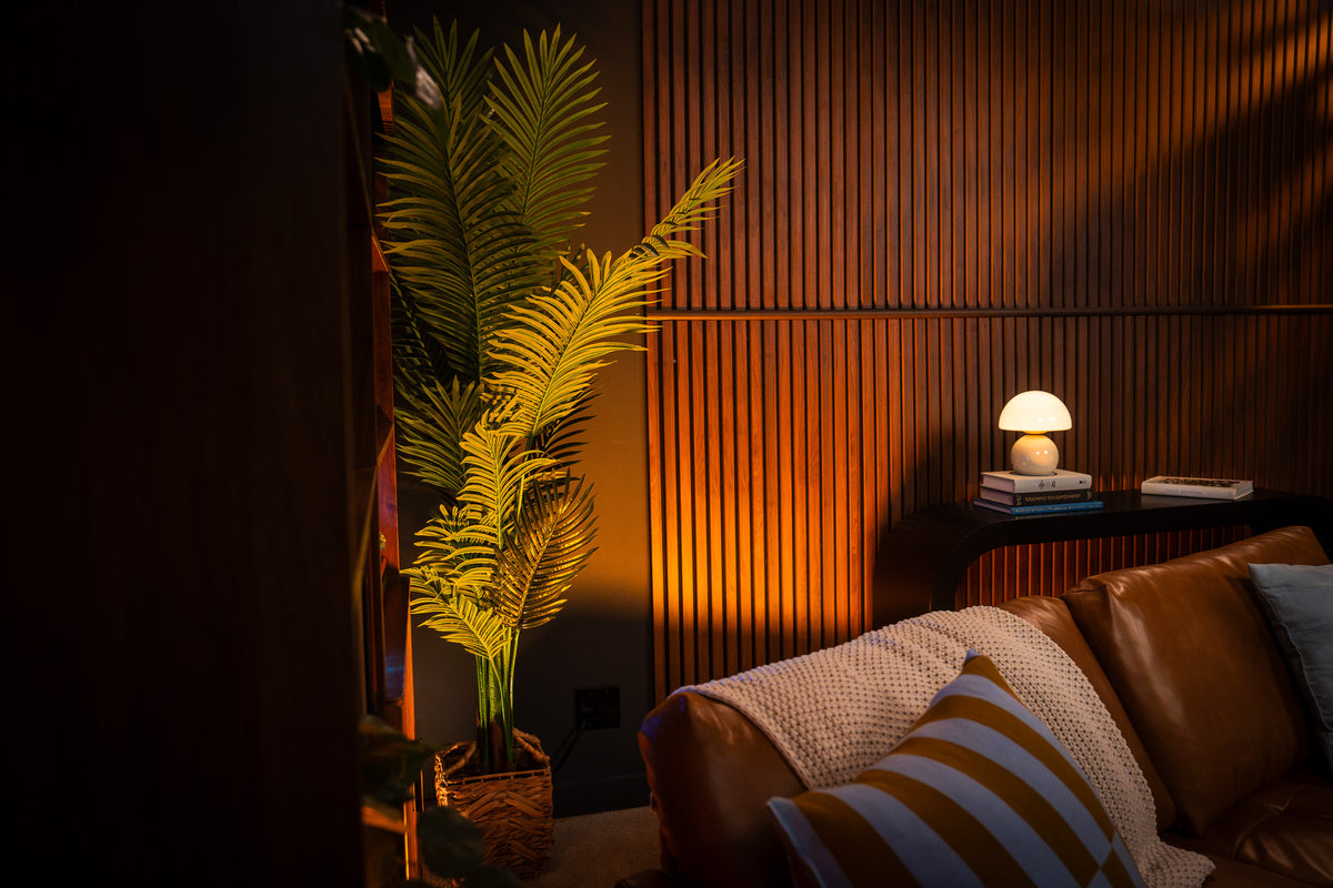 A warm, cozy living room corner with a brown leather sofa, striped pillows, a lit table lamp on a side table, and a tall artificial palm plant against a slatted wood wall.
