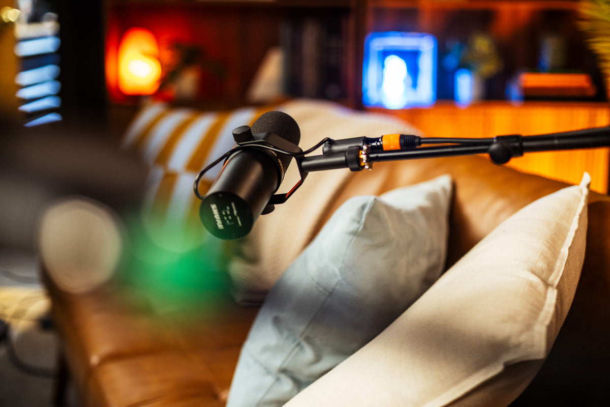 A black studio microphone mounted on an adjustable boom arm, shown in a softly blurred living room setting with a brown leather sofa, cream pillows, and warm ambient lighting in the background.