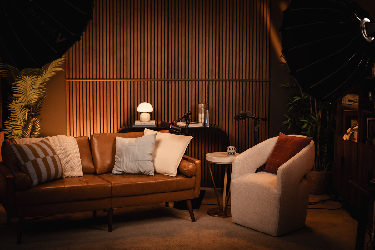 A warm, moody podcast studio setup featuring a brown leather sofa with striped and cream pillows, a cream armchair with a rust pillow, and a slatted wood accent wall in the background.