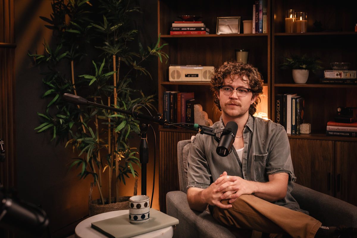 A man with curly hair and glasses sits in a gray armchair in a cozy, dimly lit room with bookshelves and a plant, speaking into a microphone mounted on a stand.