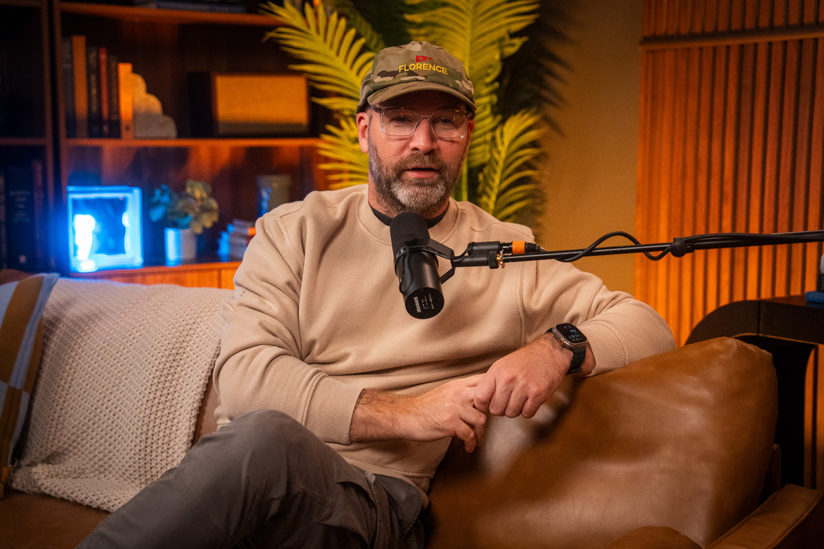 A man wearing a beige sweatshirt, gray pants, and a camouflage cap sits on a brown leather couch in a cozy room, speaking into a black microphone mounted on a boom arm.