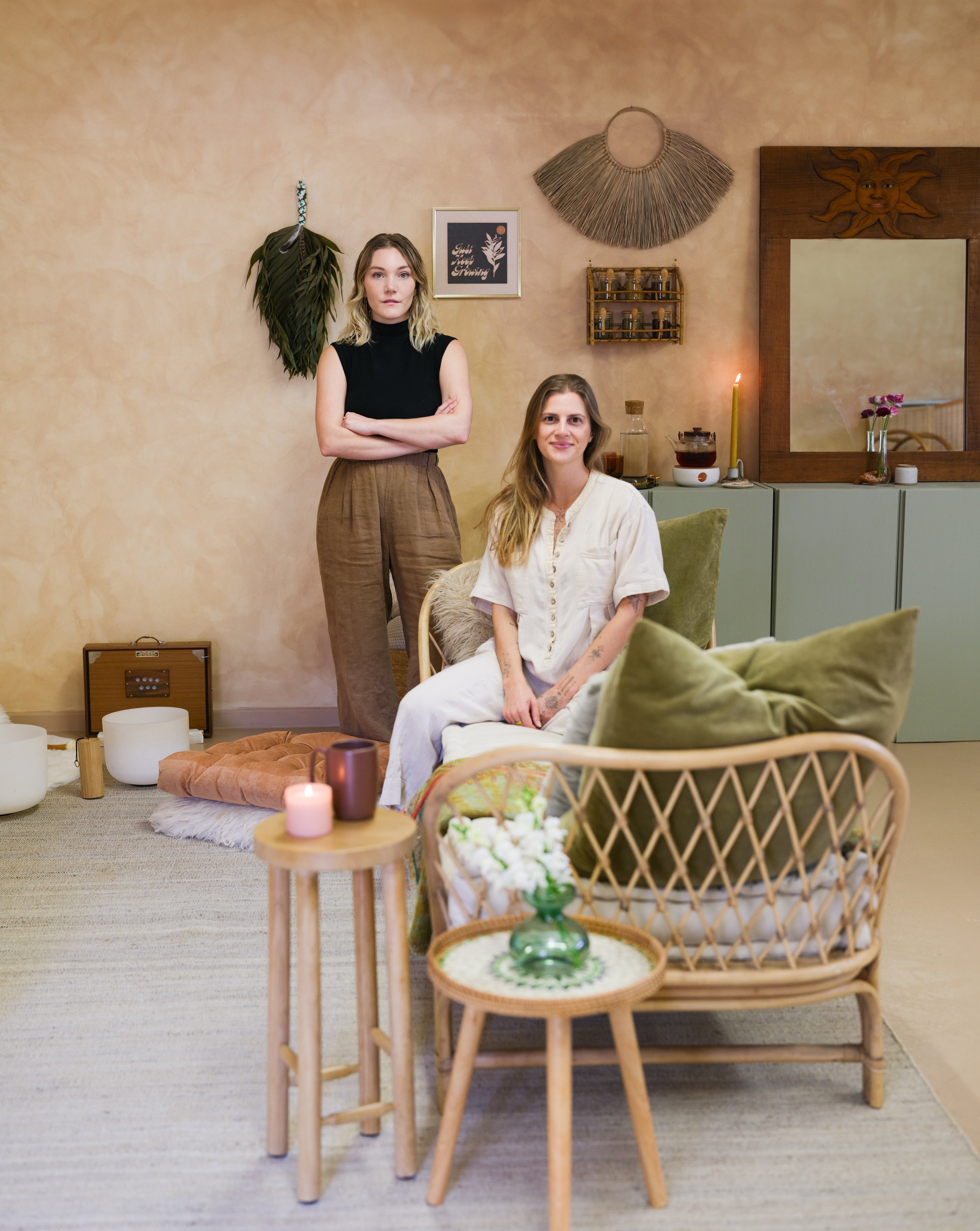 Two women posing in a beautifully styled bohemian tattoo studio. One woman stands on the left wearing a black sleeveless top and brown trousers, while the other sits on a rattan armchair in the foreground wearing a cream button-up shirt and white trousers.