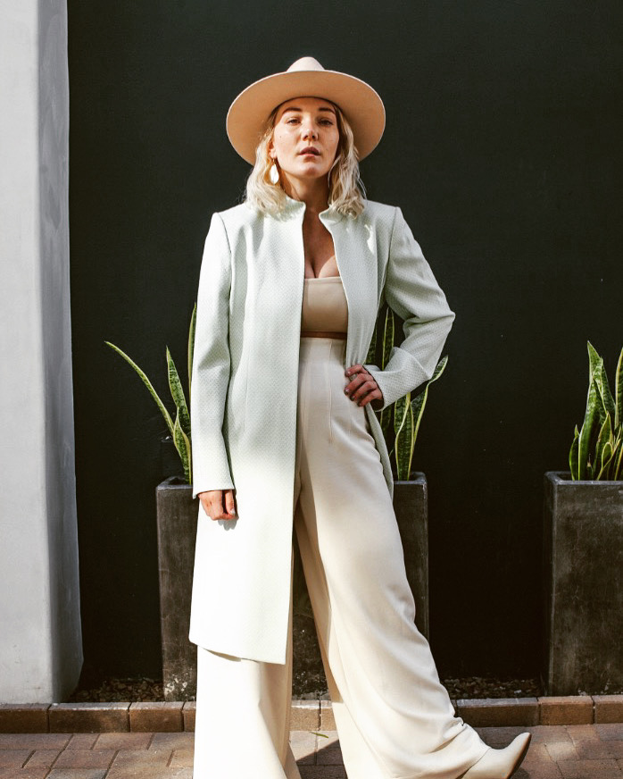 A woman stands outdoors wearing a light sage green long-line blazer over a beige top and wide-leg cream trousers, with a wide-brimmed beige hat. She is posed in front of a dark wall with potted plants.