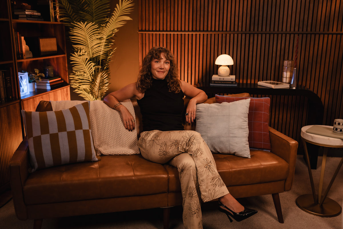 Head designer sits on a brown leather sofa in a warm, wood-paneled room, wearing a black sleeveless top and beige patterned pants with black heels. The sofa has decorative pillows, including a striped pillow and a light blue pillow.