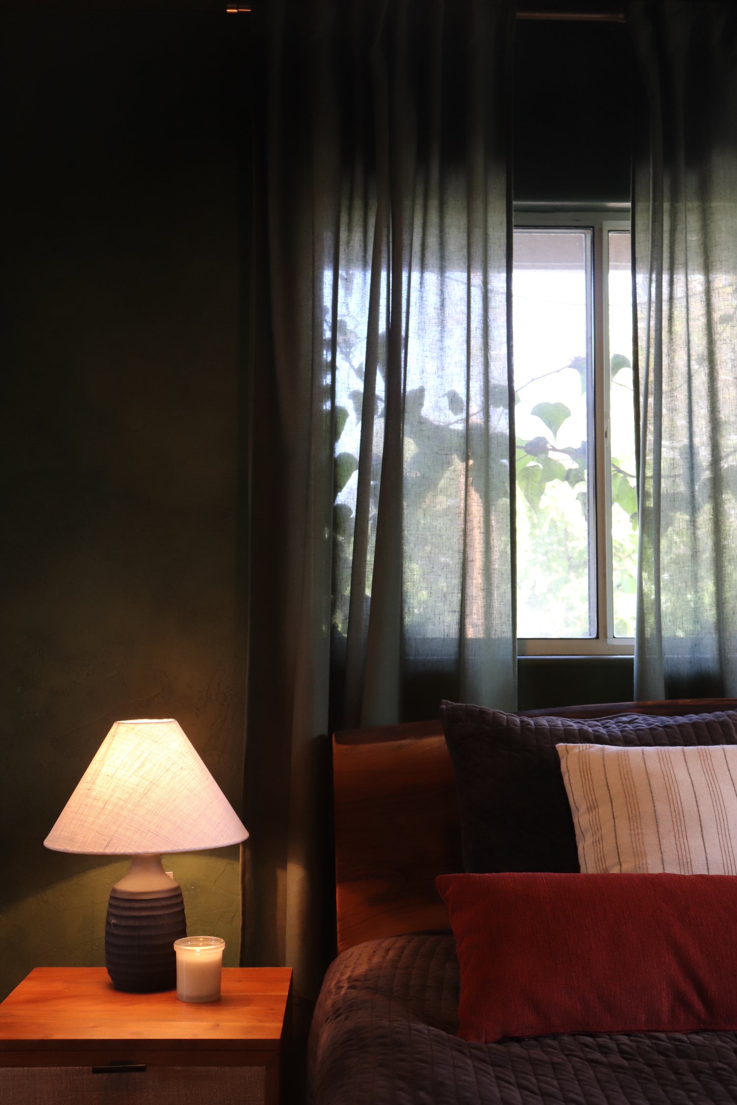 A dimly lit bedroom corner showing a bed with dark bedding, a lit bedside lamp on a wooden nightstand, and a window with sheer curtains in the background.
