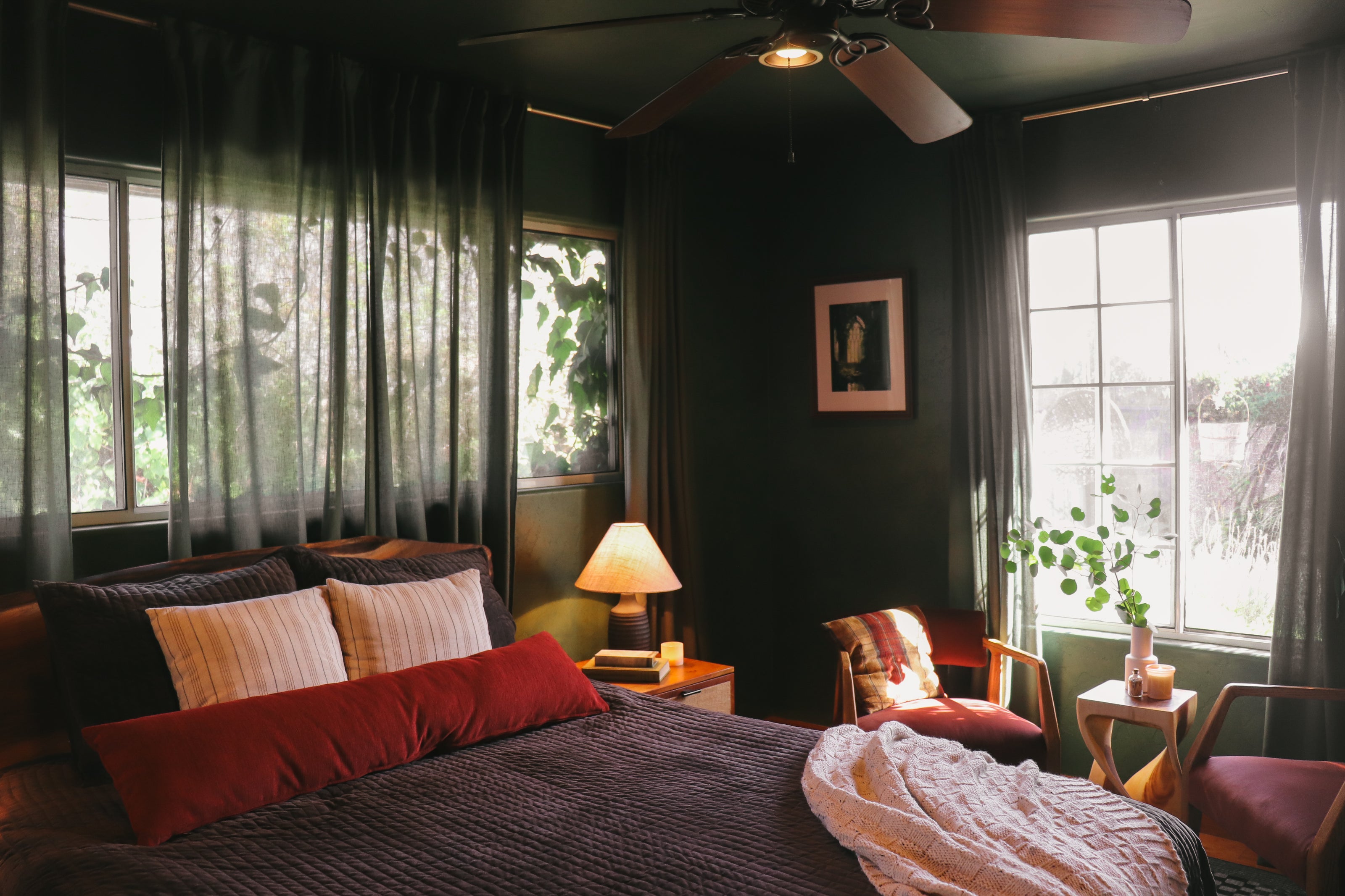 A cozy bedroom interior with a bed dressed in dark textured bedding and red pillows, dark green walls, sheer curtains, a ceiling fan, bedside lamp, and matching armchairs by large windows.