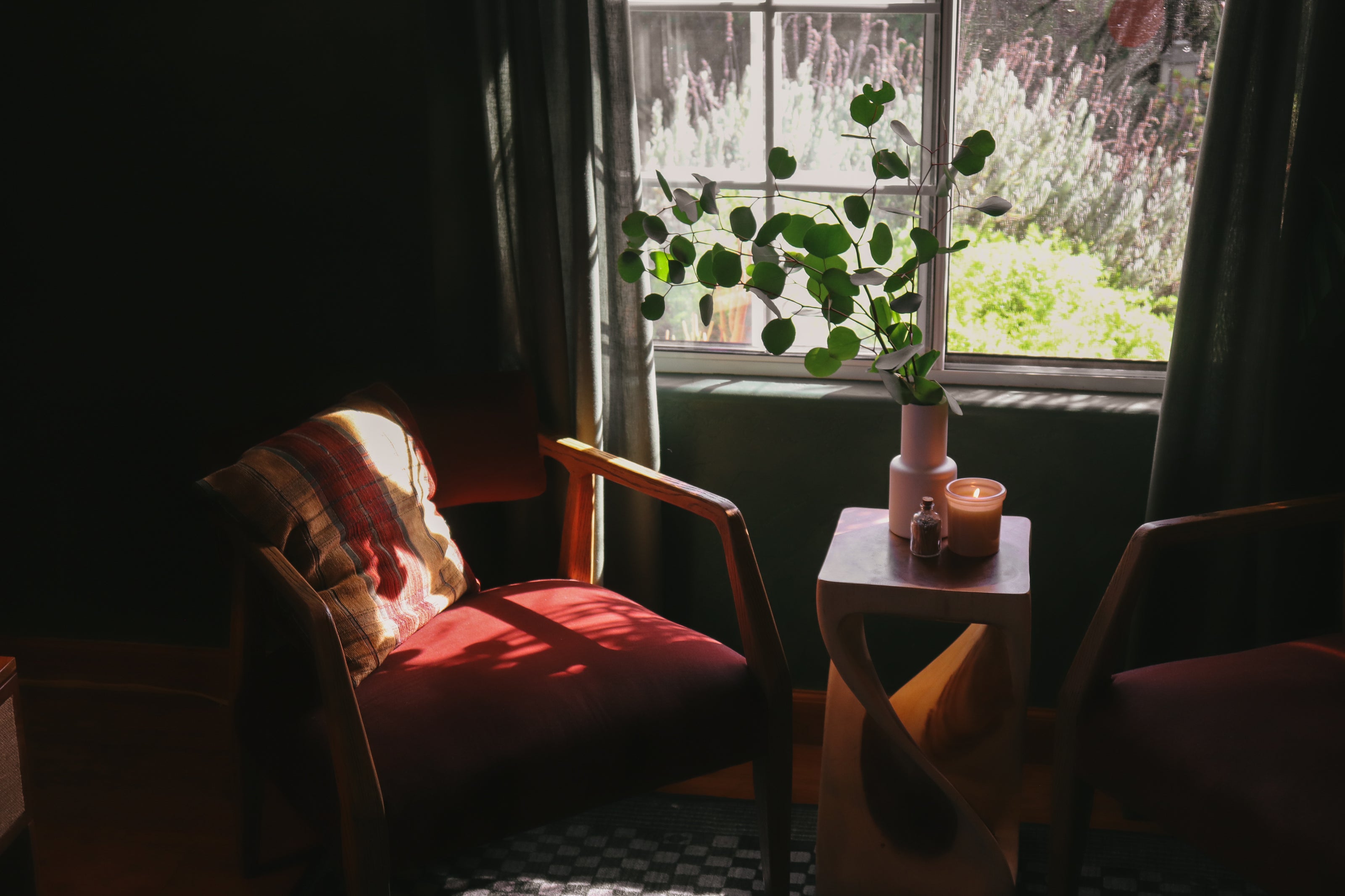 A cozy interior scene with a wooden armchair in warm sunlight next to a small side table holding a vase with green leaves and candles, set in front of a window with dark curtains.