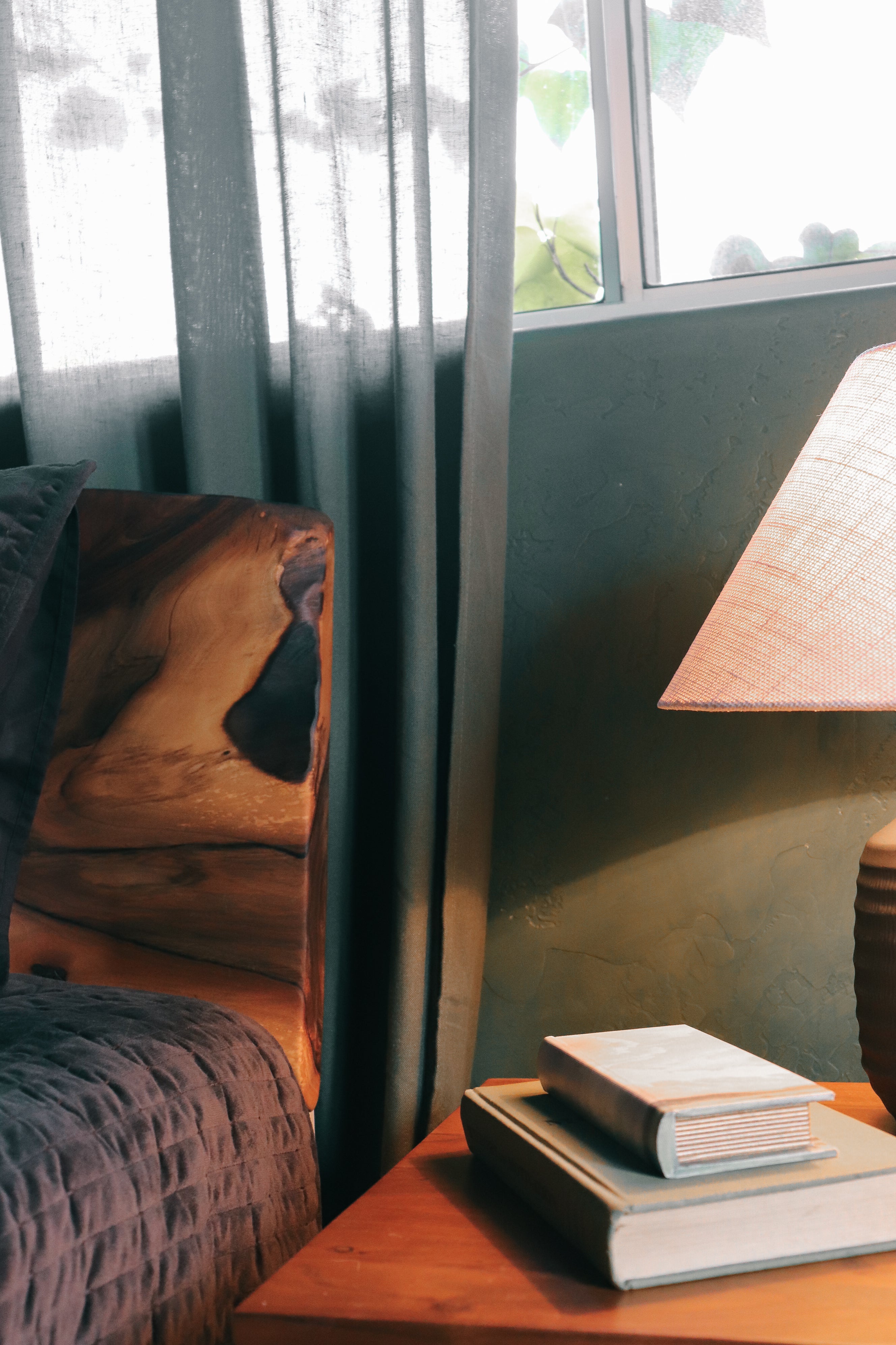 A cozy interior corner with a dark wood armchair, sheer gray curtains, a lit table lamp on a wooden side table, and stacked books near a window.