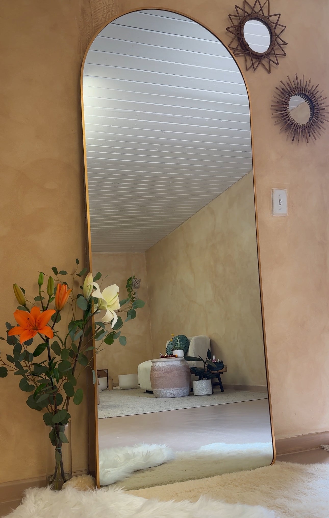 An arched full-length floor mirror with a gold-toned frame leaning against a beige wall, with a reflection of a room behind it. A decorative floral arrangement in a vase and two round wall mirrors are visible on the left and right sides of the frame.