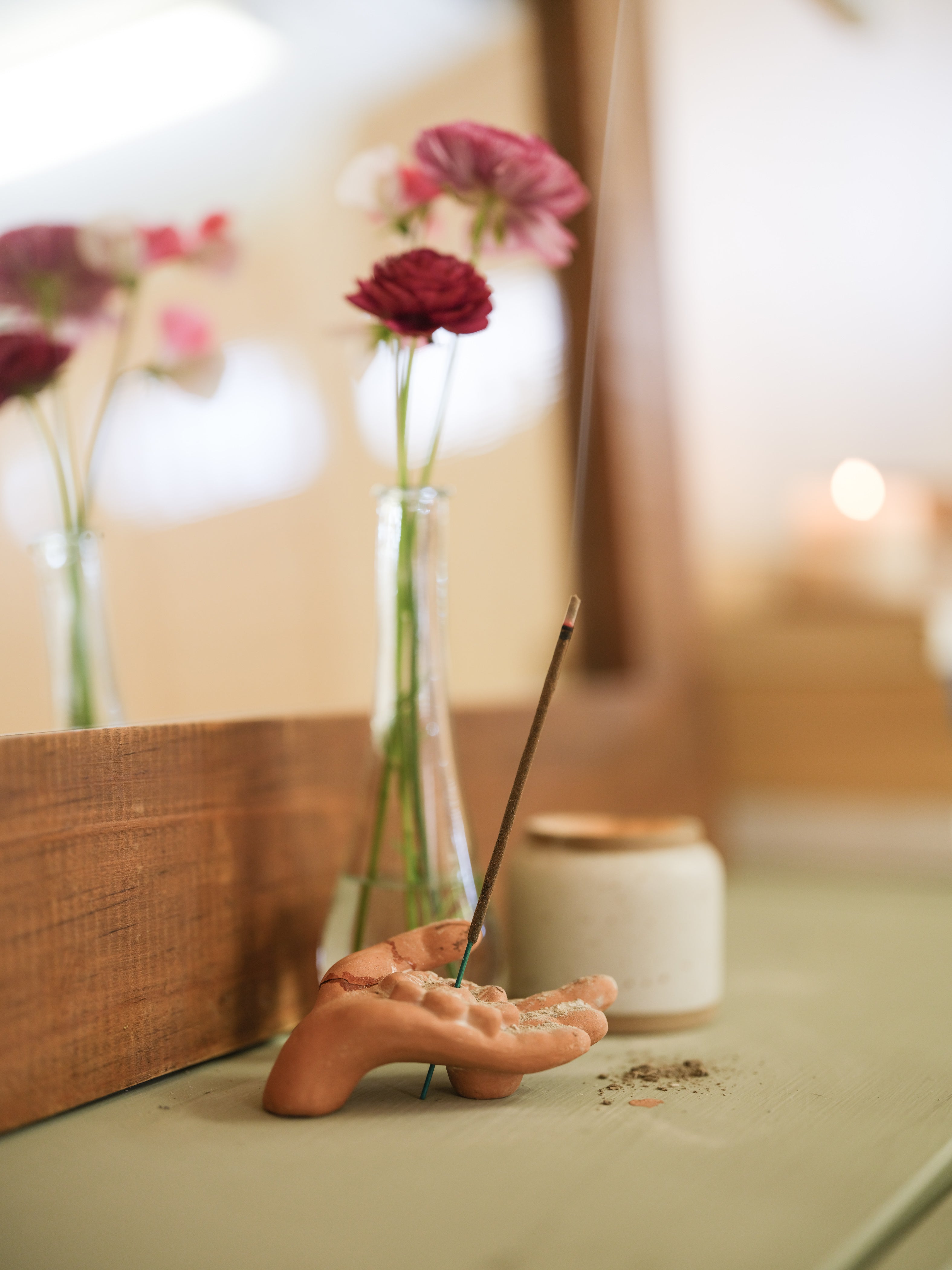 A decorative hand-shaped incense holder with a burning incense stick, set on a tabletop beside a small jar and glass vases with pink flowers in a softly lit indoor setting.