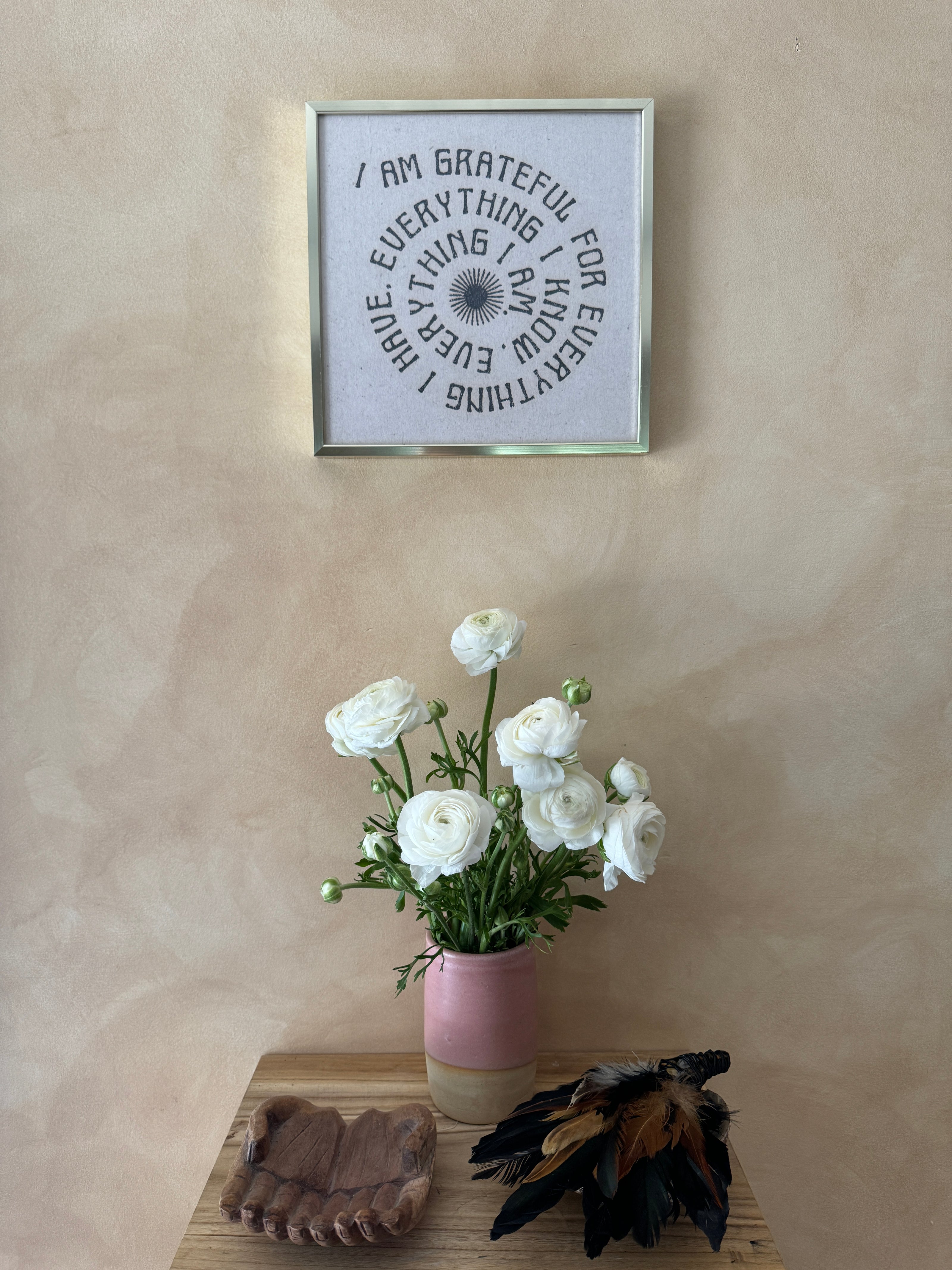 A framed spiral quote print with black text on a white background hangs on a textured beige wall above a wooden table styled with white ranunculus flowers in a pink vase, a wooden tray, and dried feathers.