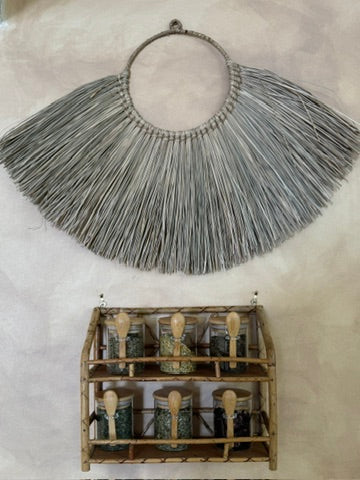 A large woven straw collar necklace with a circular top band, displayed above a small bamboo spice rack filled with jars on a textured off-white background.