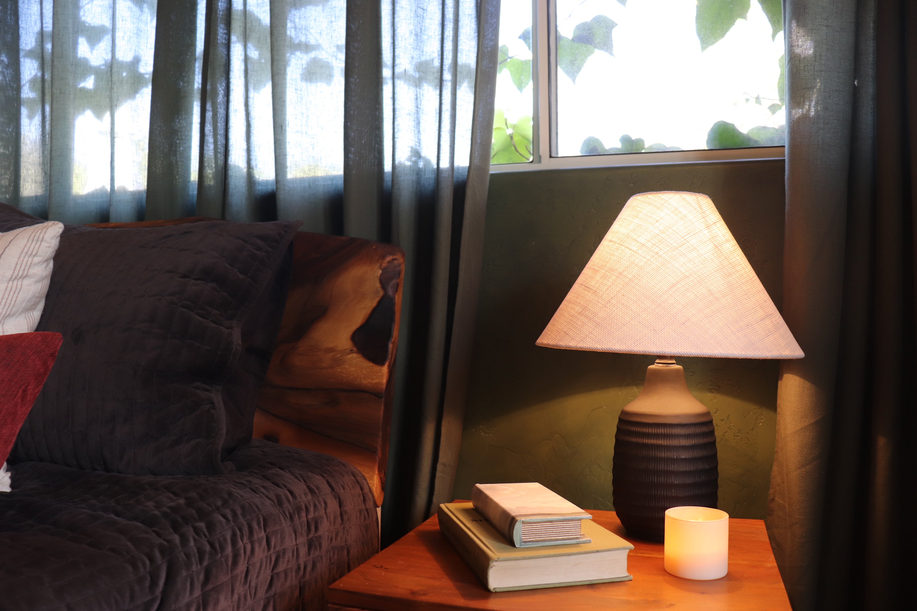 A lit table lamp with a textured beige shade and dark ribbed base sits on a wooden nightstand beside a couch, with a candle and stacked books in front of a window with sheer curtains.