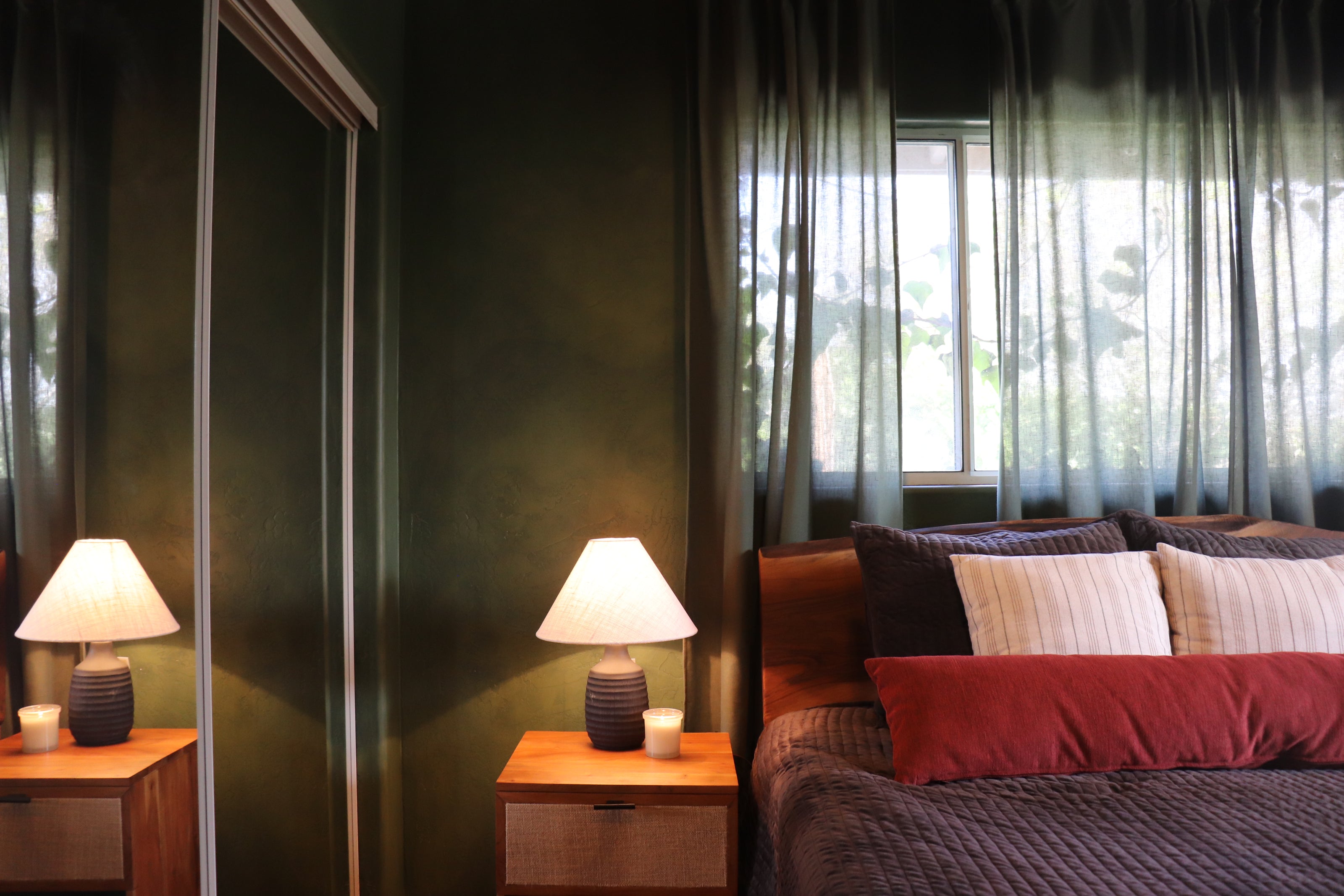 A styled bedroom interior with dark teal walls, sheer gray curtains, two wooden nightstands with lit table lamps, and a bed with dark gray bedding and red and cream pillows.