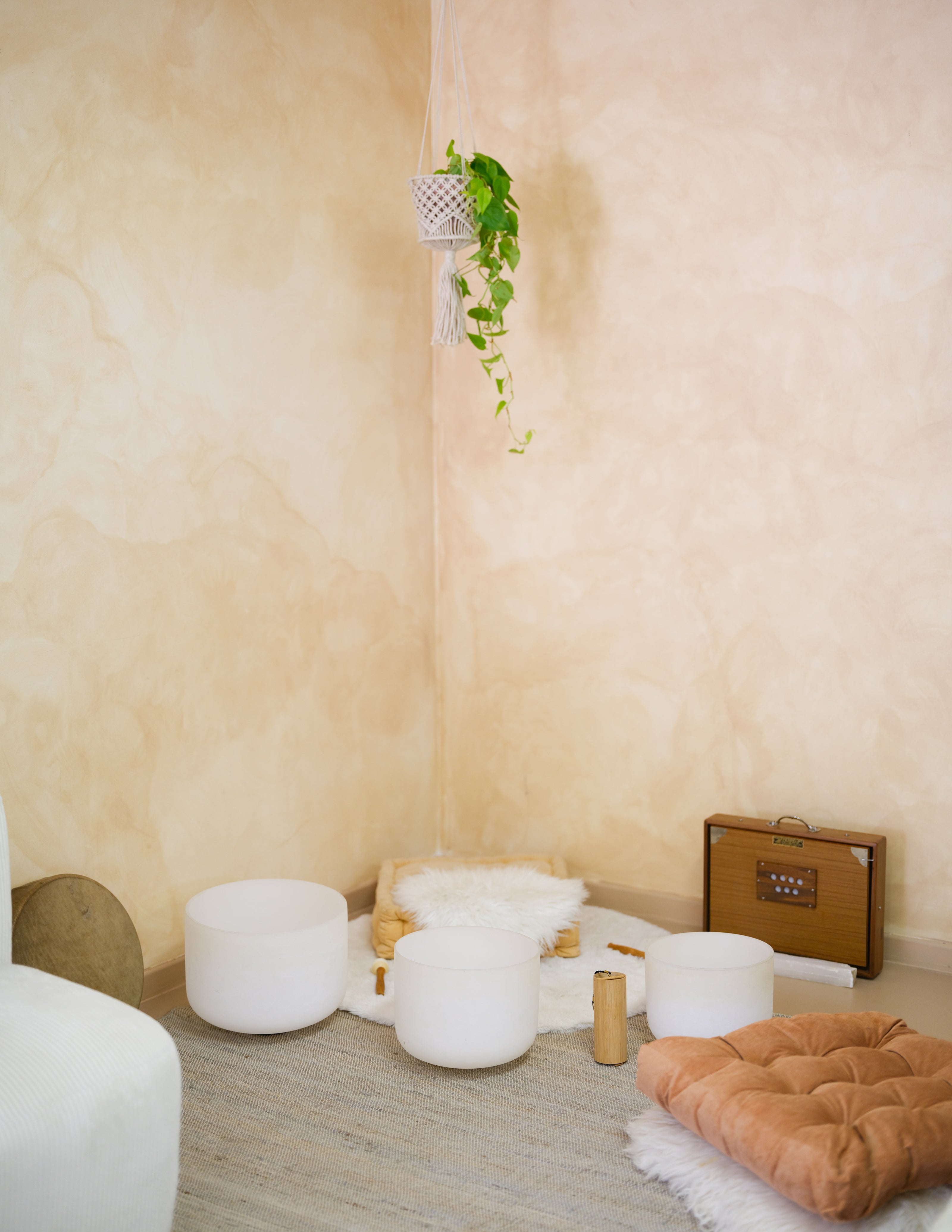 A styled corner of a room with textured beige plaster walls and a gray floor, featuring several white cylindrical bowls, a small brown suitcase, a peach cushion on a white rug, and a hanging green plant.