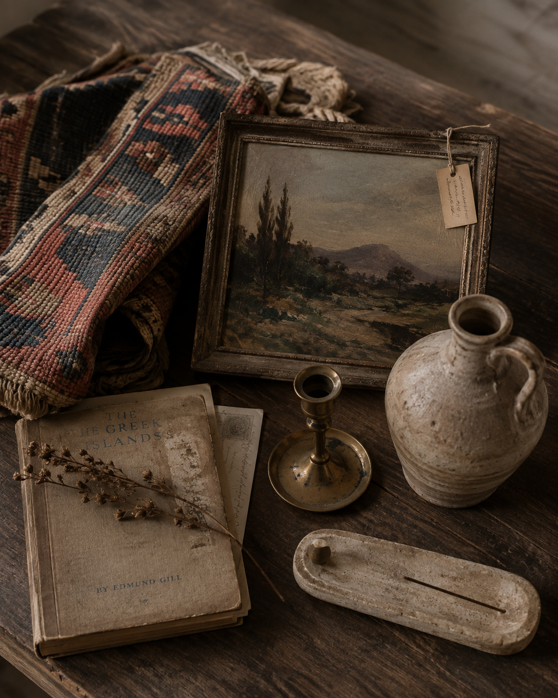 A styled flat-lay of vintage home decor items on a dark wooden table, including a small landscape painting in an ornate frame, a rustic ceramic jug, a brass candlestick, a weathered book, a patterned textile, and a wooden tray.
