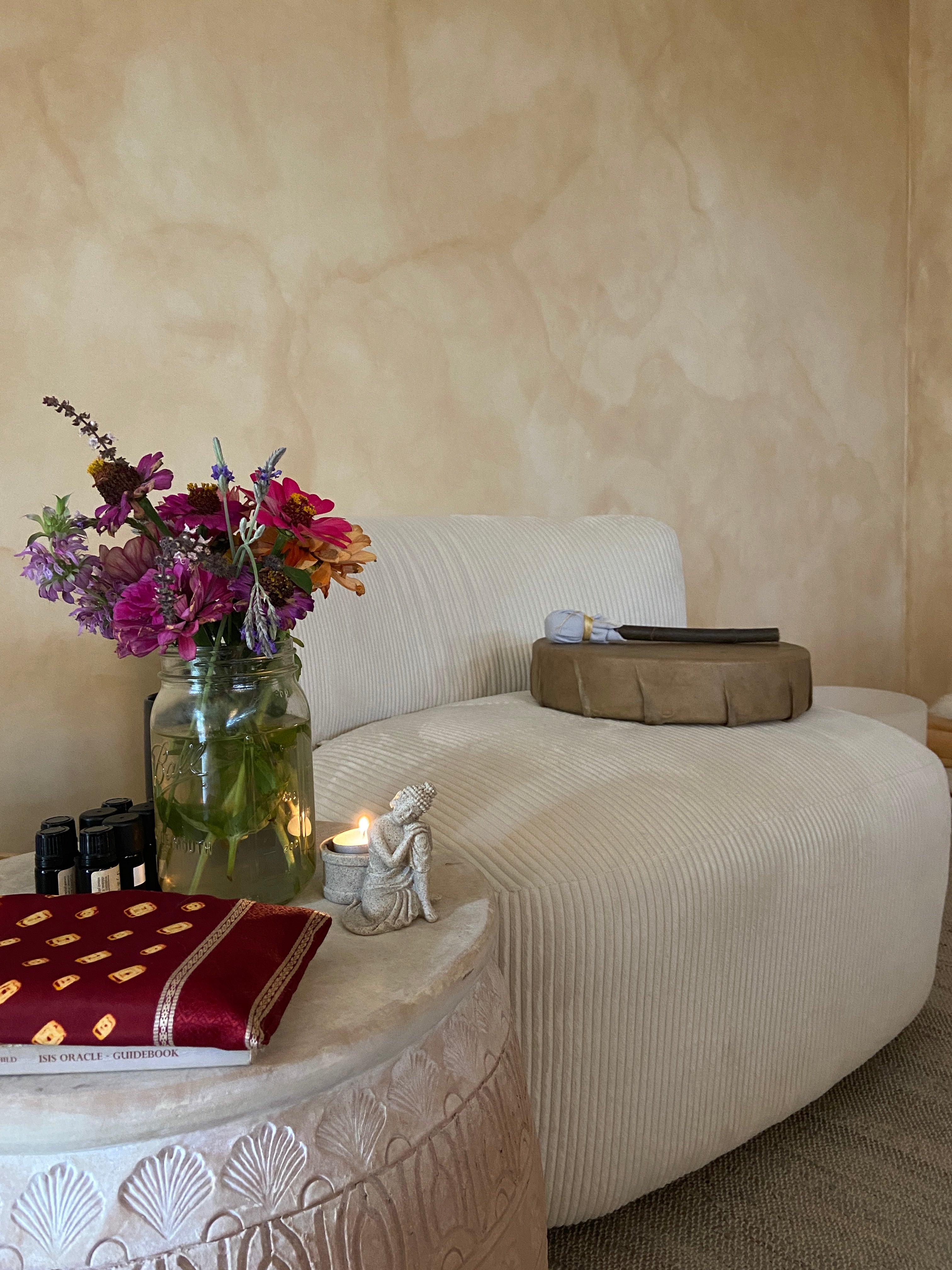 A close-up, angled view of a beige upholstered sofa with textured and ribbed cushions, decorated with a small vase of pink and purple flowers, a lit candle, and a red decorative book.