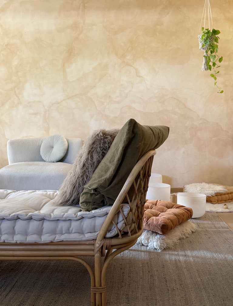 A styled interior scene showing a rattan daybed with white quilted cushions and layered pillows, including an olive green throw and a gray faux-fur pillow, set against a warm beige plaster wall.